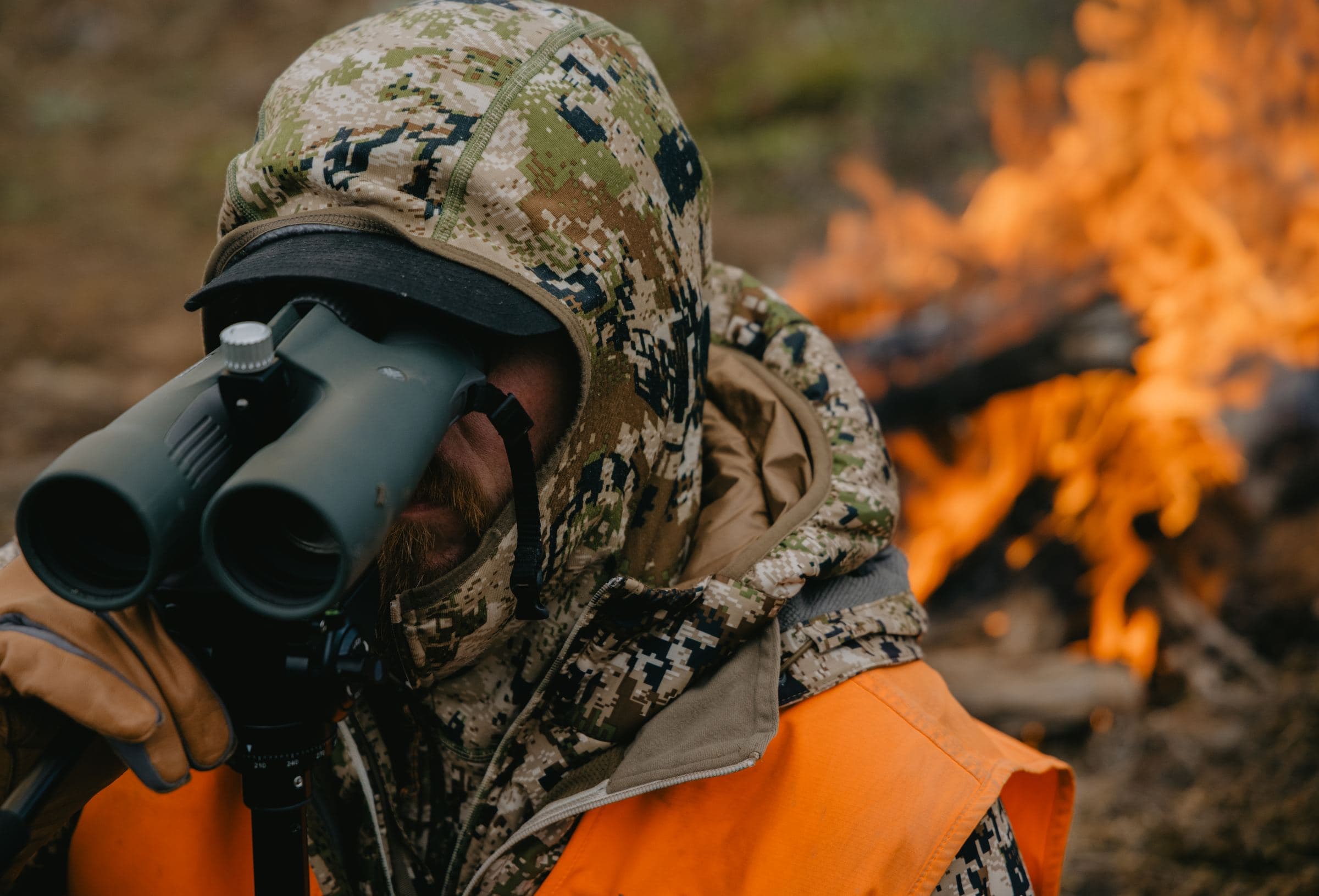 Close-up of a hooded hunter glassing on a tripod beside a campfire