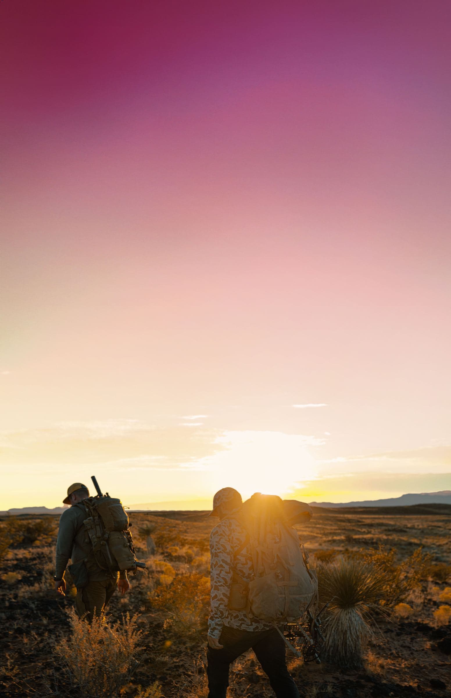 Two hunters walking at sunset under an intense magenta-to-yellow sky