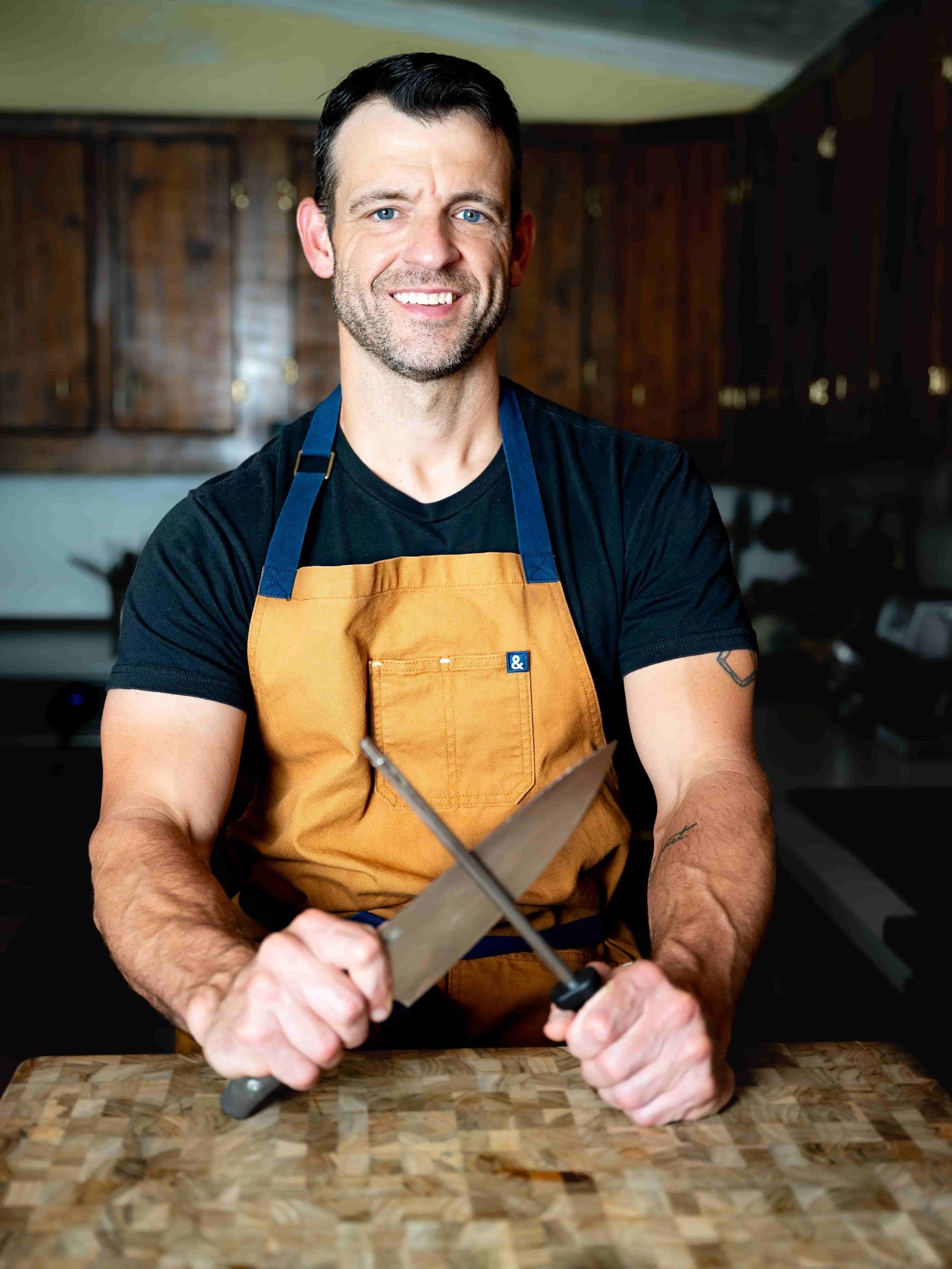 Posed indoor portrait of a man in an apron holding a knife and steel