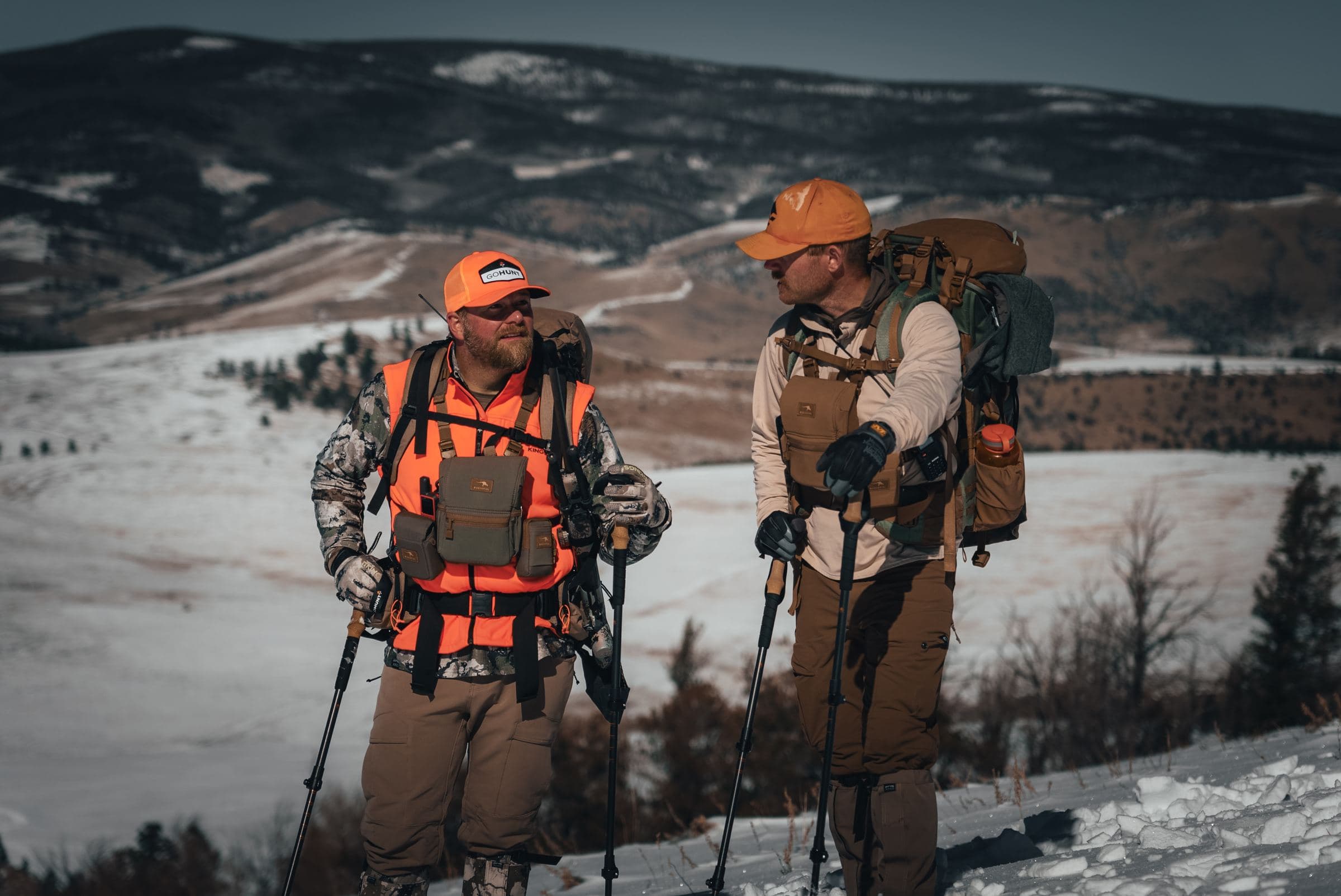 Two elk hunters in blaze orange with packs pause on a snowy hillside