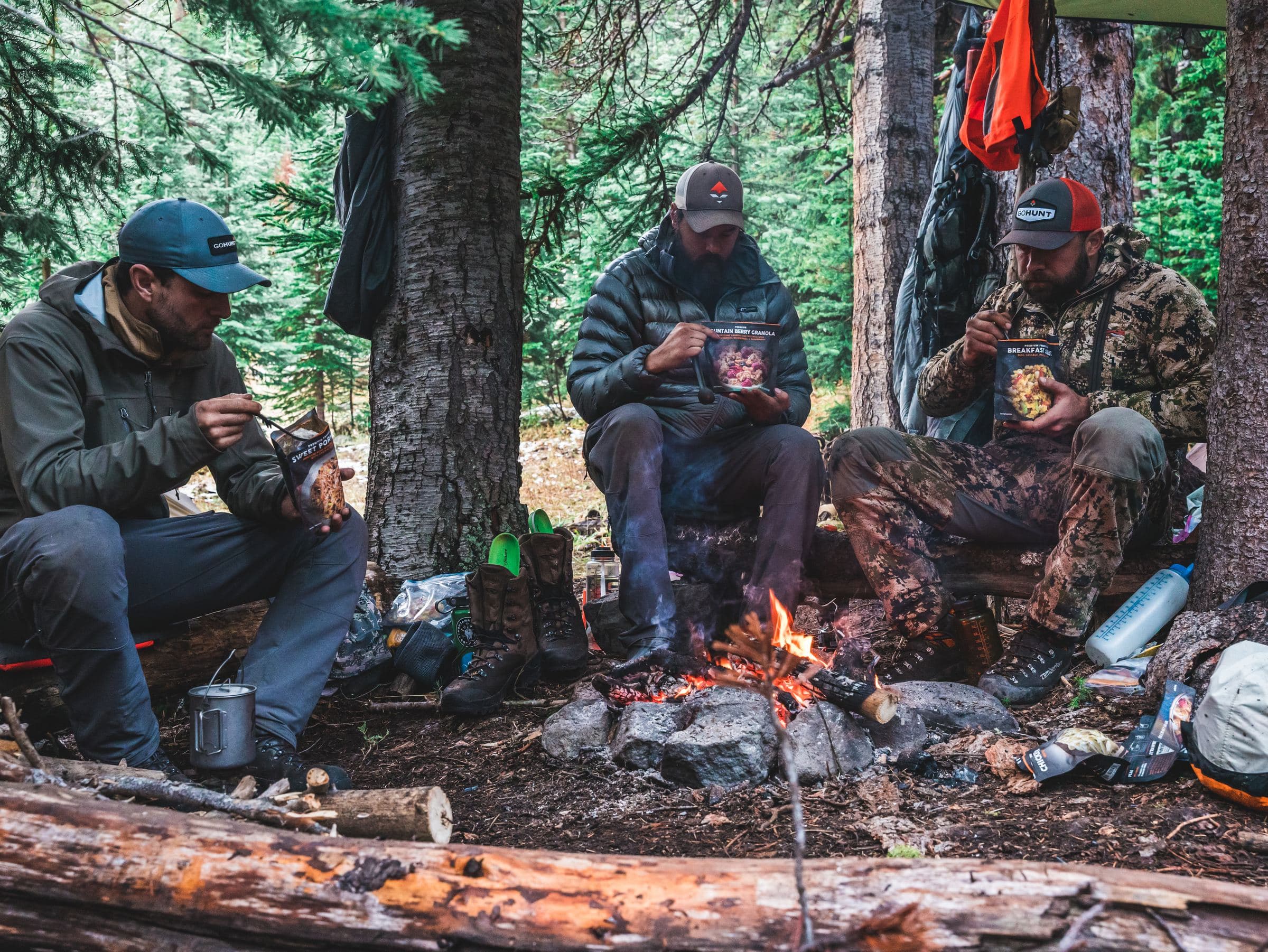 Three hunters eat around a small campfire in the timber