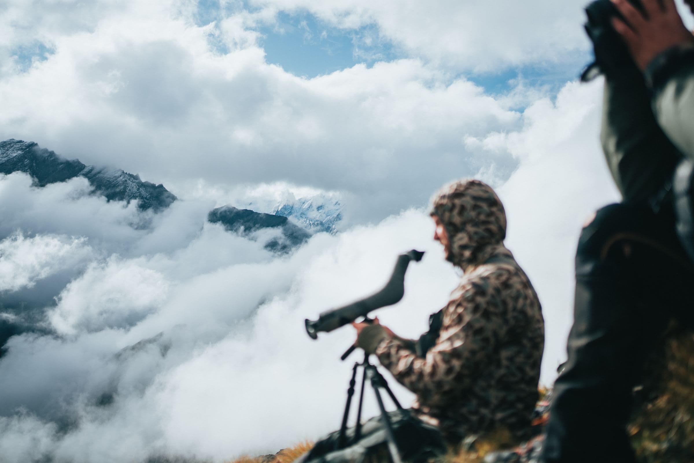 Hunter behind a spotting scope on a ridge above a sea of clouds